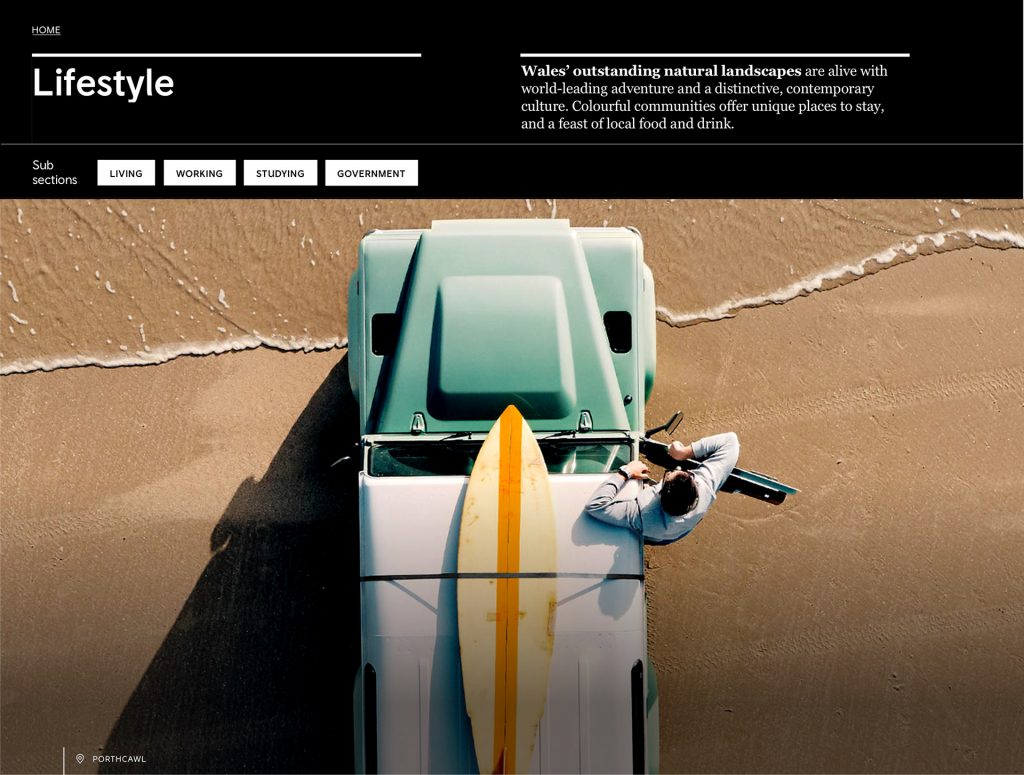 Shows the heading of an article for wales.com and visitwales.com. The text is places at the top followed by tags in the next row. Below those two elements there's a large image with a man coming out of a jeep in the beach. The jeep has a surfboard on the roof. 